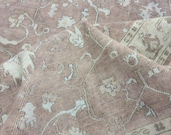 May include: A dusty rose area rug featuring a repeating pattern of off-white and light gray floral and geometric motifs. The rug has a visible woven texture with a folded corner revealing the underside, showcasing the intricate, symmetrical design.