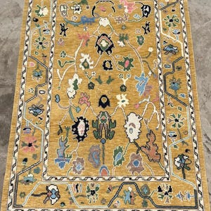 May include: A rectangular area rug with a mustard yellow background featuring a symmetrical, intricate pattern of stylized floral and organic shapes in shades of blue, green, pink, white, and black, framed by a geometric border and white fringe.