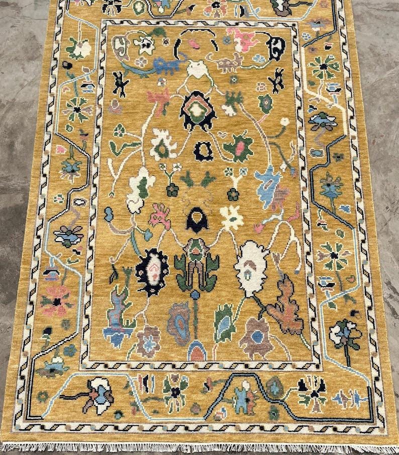 May include: A rectangular area rug with a mustard yellow background featuring a symmetrical, intricate pattern of stylized floral and organic shapes in shades of blue, green, pink, white, and black, framed by a geometric border and white fringe.