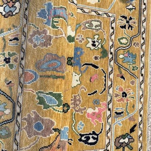 May include: A mustard yellow area rug featuring a repeating floral pattern in shades of blue, pink, green, and white. The rug has a distinct border with a geometric zigzag design and a central field filled with stylized botanical motifs.