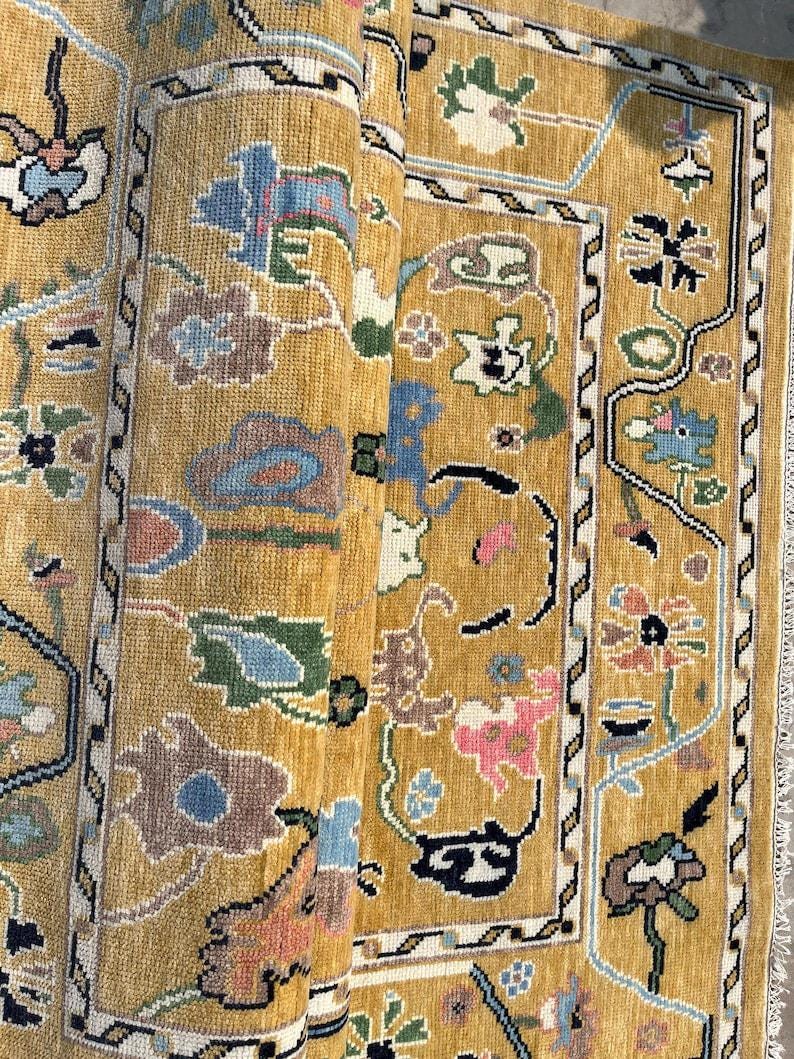 May include: A mustard yellow area rug featuring a repeating floral pattern in shades of blue, pink, green, and white. The rug has a distinct border with a geometric zigzag design and a central field filled with stylized botanical motifs.