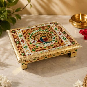 May include: A rectangular, low-profile stand featuring a central circular medallion with a peacock motif. The surface is decorated with intricate gold, red, green, and white enamel patterns. It is supported by four short, ornate, gold-toned legs.