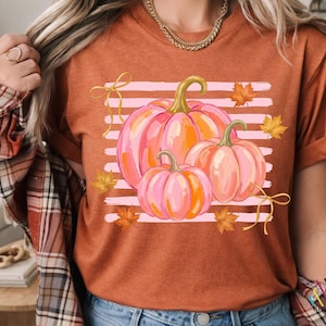 May include: A burnt orange t-shirt featuring a graphic print of three pink and orange pumpkins centered over horizontal light pink stripes. The design includes scattered golden yellow maple leaves and two delicate yellow ribbon bows.