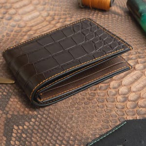 Può includere: Un portafoglio bifold marrone scuro caratterizzato da una texture esterna con motivo a pelle di coccodrillo. Il portafoglio è realizzato con cuciture arancioni visibili lungo i bordi e presenta una fodera interna liscia di colore marrone scuro.