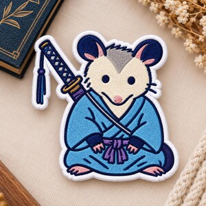 May include: An embroidered patch featuring a cream-colored opossum wearing a blue kimono with a purple sash. The opossum has a katana strapped to its back, complete with a blue hilt and a matching tassel. The design is outlined in white thread.