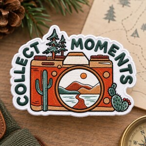 May include: An embroidered patch featuring an orange camera with the text COLLECT MOMENTS in dark green. The camera lens displays a desert landscape with mountains, a sun, and a river. A green cactus and two pine trees are also depicted.