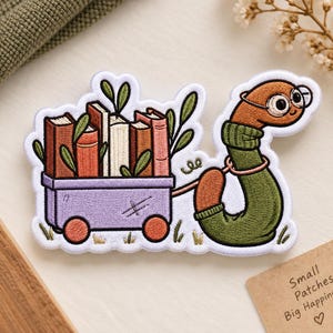 May include: An embroidered patch featuring a brown worm wearing round glasses and a green sweater, pulling a purple wagon filled with five upright books and green leaves. The patch has a white border and a detailed, multi-colored thread design.
