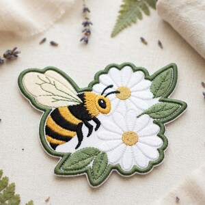 May include: An embroidered patch featuring a bumblebee with yellow and black stripes and light cream wings, positioned next to two white daisies with yellow centers and green leaves, all outlined with a dark green scalloped border.