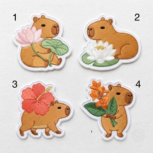 May include: Four embroidered patches featuring brown capybaras with various flowers. Patch 1 shows a capybara with a pink lotus, 2 with a white lotus, 3 with a large red hibiscus, and 4 with orange flowers. Each patch has a white border.