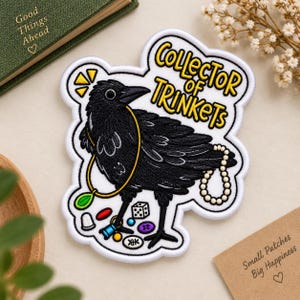 May include: An embroidered patch featuring a black crow with a yellow necklace, a string of white pearls, and various small trinkets like a die, thimble, and buttons. The text reads Collector of Trinkets in yellow letters on a white background.