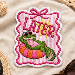 May include: An embroidered patch featuring a green alligator wearing heart-shaped sunglasses, lounging on a pink and orange striped pool float. The background is white with pink vertical stripes, a yellow star, and the word LATER in pink text.