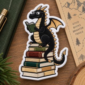 May include: An embroidered patch featuring a black dragon with gold wings and horns wearing glasses while reading a small green book. The dragon is perched atop a stack of five books in various colors including green, red, and tan.