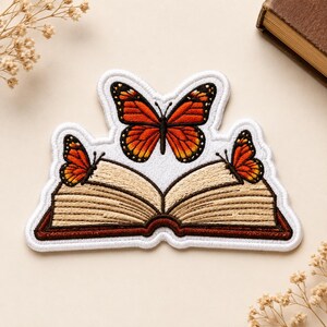 May include: An embroidered patch featuring an open book with tan pages and a dark brown cover. Two orange and black monarch butterflies are perched on the open pages, while a third, larger monarch butterfly is positioned above the book.