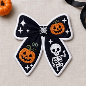 May include: A black fabric bow with white embroidered edges featuring a central spiderweb, two orange jack-o'-lanterns, a white skeleton, and several four-pointed star shapes. The bow has a textured surface and is shaped like a decorative accessory.