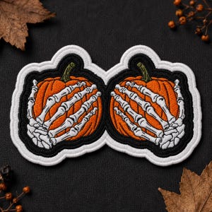May include: An embroidered patch featuring two orange pumpkins, each being held by a white skeleton hand. The design is outlined in black and white thread, creating a cohesive shape with a dark border that highlights the skeletal fingers.