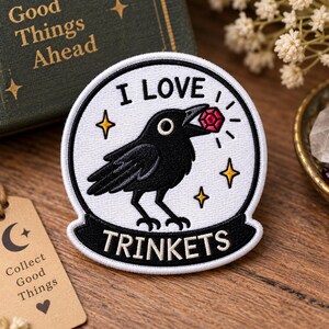 May include: A circular embroidered patch featuring a black crow holding a red faceted gem in its beak. The white background includes the text I LOVE TRINKETS in black, surrounded by yellow star icons and a black border with a banner at the bottom.