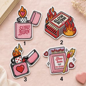 May include: Four embroidered patches featuring fire and heart motifs. Patch 1 is a pink lighter with the text SLOW BURN. Patch 2 is a matchbox with the text SLOW BURN and flames. Patch 3 is a pink lighter with a heart. Patch 4 is a matchbox with the text Slow burn MADE FOR READERS.