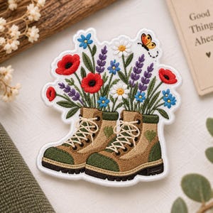 May include: An embroidered patch featuring a pair of tan and green hiking boots filled with a bouquet of red poppies, white daisies, purple lavender, and blue wildflowers. A small orange butterfly is embroidered near the top right corner.