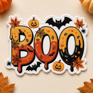 May include: An embroidered patch featuring the word BOO in a dripping, orange and black font. The design includes orange pumpkins, black bats, spider webs, small spiders, and orange maple leaves on a white background with a white border.
