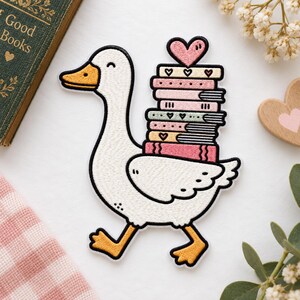 May include: An embroidered patch featuring a white goose with an orange beak and feet, walking while carrying a stack of six colorful books on its back, topped with a small pink heart. The books are decorated with various heart and line patterns.