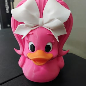 May include: A bright pink rubber duck featuring a large white bow attached to the top of its head. The duck has a vibrant orange beak, black eyes with white pupils, and sculpted pink hair that flares out on both sides of its head.