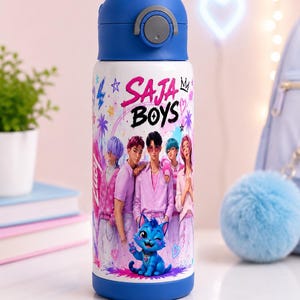 Könnte beinhalten: Eine weiße Trinkflasche mit blauem Deckel und Boden, bedruckt mit einer Grafik von fünf Personen in rosa Outfits und einer blauen Katze. Der Text SAJA BOYS ist in Pink und Schwarz gedruckt, ergänzt durch Sterne, Blitze und Palmenmotive.