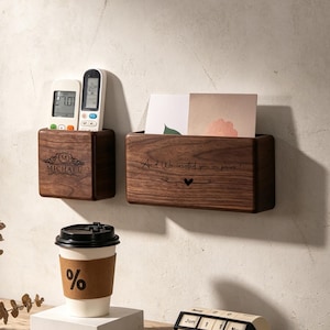 Puede incluir: Dos organizadores de pared rectangulares de madera oscura. El más pequeño está grabado con 'MICHAEL' y contiene dos mandos a distancia. El más grande está grabado con 'And We created you in pairs' y un corazón, conteniendo dos tarjetas.
