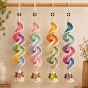 May include: Four hanging spiral crochet ornaments in vibrant pastel color gradients. Each piece features a twisted design, a wooden bead, and a bottom tassel adorned with a small crocheted flower and green leaves, suspended from a wooden rod.