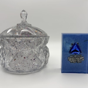 Peut inclure: Un pot en résine transparent à facettes avec couvercle, rempli de paillettes multicolores, placé à côté d'un bloc rectangulaire en résine bleue orné d'une grosse pierre précieuse triangulaire bleue et d'une rangée de strass transparents.