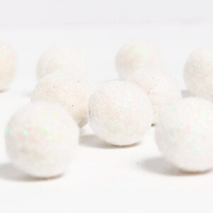 May include: A collection of white, glitter-covered spheres scattered on a white surface. The spheres have a soft, textured appearance with iridescent glitter accents, creating a sparkling effect. The objects appear to be decorative craft supplies.