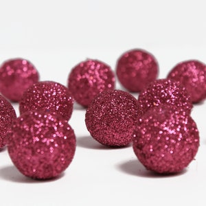 May include: A collection of glitter-covered, round decorative spheres in a deep magenta color. The spheres are scattered on a white surface, creating a festive and eye-catching display. These decorative balls are ideal for holiday or craft projects.