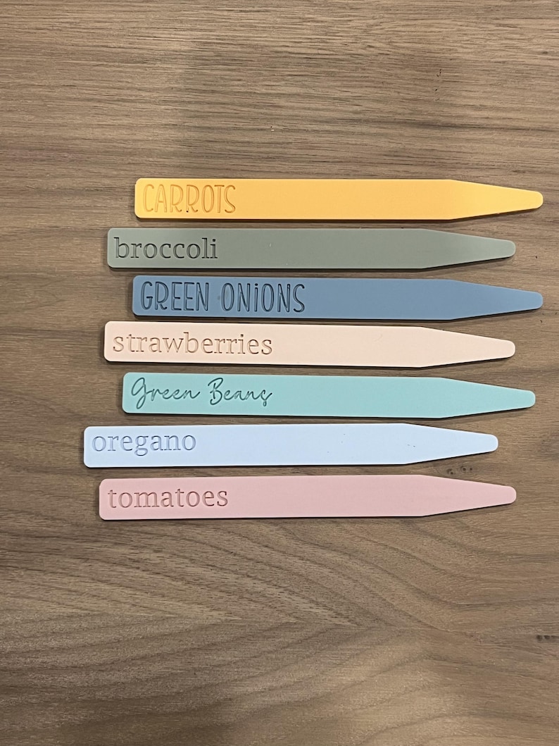 May include: Seven colorful plant markers with different names written on them: Carrots, broccoli, Green Onions, strawberries, Green Beans, oregano, and tomatoes.