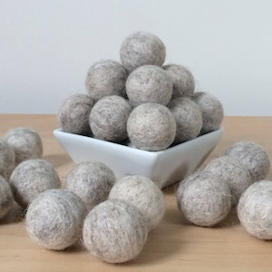 May include: A pile of gray felt balls in a white square bowl, with more gray felt balls scattered around the bowl on a light wood surface.