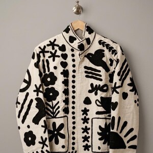 Puede incluir: Una chaqueta de color crema con motivos bordados en negro que incluyen flores, corazones, un pájaro, un sol y formas geométricas. La chaqueta tiene cuello alto, mangas largas y cierre frontal de botones, colgada en una percha de madera.