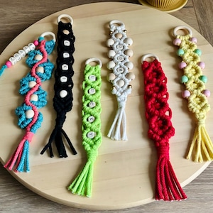 Pode incluir: Seis porta-chaves de macramé em várias cores, incluindo azul, preto, verde lima, branco, vermelho e amarelo. Cada porta-chaves tem um anel de madeira, padrões de corda atada e alguns incluem contas de madeira com as letras ZOSIA, LUKKI, LUCKY ou DOM.