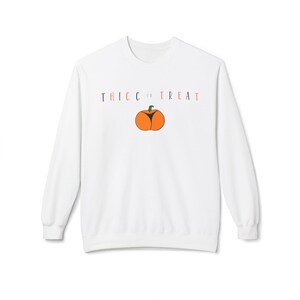 May include: A white long-sleeve sweatshirt featuring the text THICC OR TREAT in black and orange lettering. Centered below the text is a graphic of an orange pumpkin with a green stem and a black V-shaped design on its front surface.