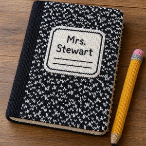 Peut inclure: Une couverture de cahier tricotée avec un motif noir et blanc et le nom "Mrs. Stewart" sur une étiquette. Un crayon jaune tricoté avec une gomme rose repose à côté du cahier. Le cahier et le crayon sont sur une surface en bois.