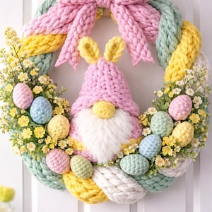 以下が含まれることがあります： パステルカラーの卵と花に囲まれた、ウサギの耳を持つピンクのノームが特徴の、手作りのイースターリース。ピンク、黄色、緑、白のニット糸で作られ、上部に大きなピンクのリボンが付いています。