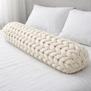 Puede incluir: Un cojín cilíndrico de color crema con un diseño de punto trenzado grueso. La almohada está colocada sobre una cama blanca con dos almohadas blancas en el fondo. La textura del punto es claramente visible.
