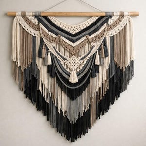 Pode incluir: Uma grande tapeçaria de macramé em tons neutros. A peça apresenta uma barra de madeira na parte superior com um padrão complexo de fibras tecidas e amarradas em tons de creme, bege, cinza e preto. Borlas decorativas pendem da parte inferior.