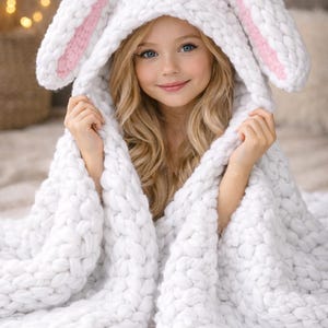 Pode incluir: Um cobertor com capuz branco de malha grossa com orelhas de coelho. As orelhas têm detalhes internos rosa. O cobertor está enrolado em uma pessoa. A pessoa está sorrindo e tem cabelo loiro e olhos azuis.