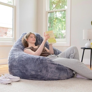 May include: A large, fluffy, blue beanbag chair. A person is sitting in the chair, reading a book. White sneakers are on the floor in front of the chair. The room has large windows and a side table with a lamp.
