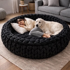 Peut inclure: Un grand lit pour animaux de compagnie rond et noir tricoté. Une personne et un chien blanc se reposent à l'intérieur du lit. Le lit est sur un tapis de couleur neutre dans un salon.