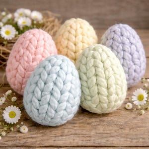 May include: A collection of six pastel-colored, knitted Easter eggs. The eggs are in shades of pink, yellow, light blue, green, and lavender. They are arranged on a rustic wooden surface, with small white flowers and greenery in the background.