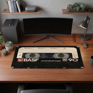 Può includere: Un tappetino da scrivania rettangolare con una grande grafica dettagliata di una cassetta BASF chromdioxid super 90 nera e beige. Il design include il testo Stereo, Mono, Dolby, Nr., BASF e 90, con due bobine di nastro visibili.