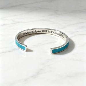 May include: A silver-toned cuff bracelet featuring a vibrant turquoise enamel inlay along the outer band. The interior surface is engraved with the black text, This too shall pass, BUT FUCKING HELL, in a clean, sans-serif font style.