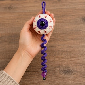 Op de afbeelding: Een gehaakt oogbolornament bestaande uit een witte bol met rode geborduurde aders, een paarse iris en een zwarte pupil. Een paarse gehaakte spiraalvormige staart hangt naar beneden en eindigt met een doorschijnende paarse en zwarte kraal.