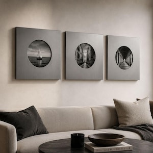 May include: Three square gray wall art panels, each featuring a circular cutout displaying a black and white photograph. The first shows a sailboat on water, the second a narrow street, and the third a long arched corridor with columns.