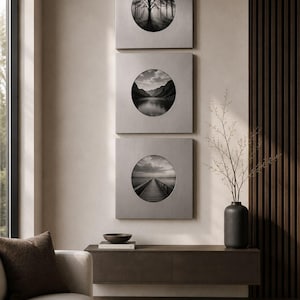 May include: Three square wall art pieces arranged vertically, each featuring a circular cutout displaying a black and white landscape photograph. The frames have a brushed metallic finish, showcasing a forest, mountain lake, and a wooden pier.