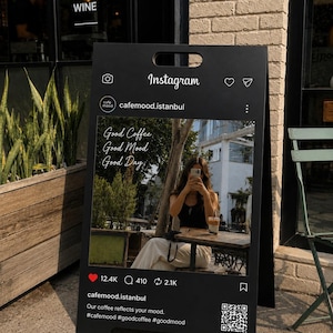 Puede incluir: Un letrero de acera negro en forma de A con una interfaz de publicación de Instagram impresa. El gráfico muestra el nombre cafemood.istanbul, una foto de una persona en una mesa, el texto Good Coffee Good Mood Good Day y un código QR.